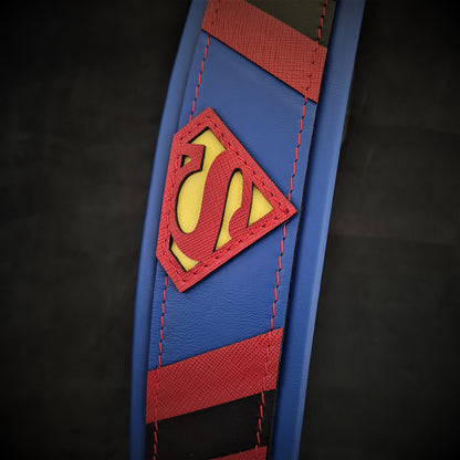 A close-up of the Superdog Collar, a blue and red strong dog collar featuring a yellow and red Superman logo patch stitched onto it, set against a dark background.